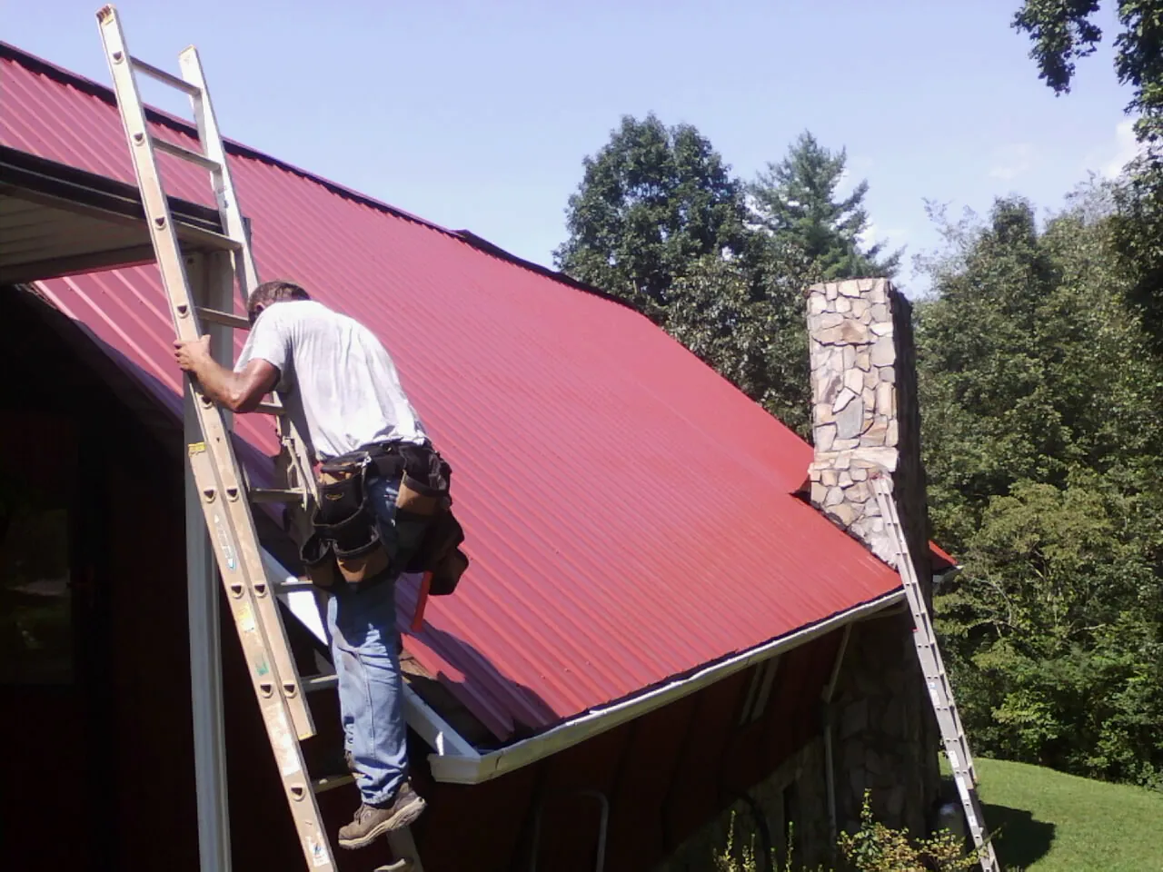 Roofer on a ladder beside a red metal roof and stone chimney — Soffit & Fascia Repair by Roofing Brothers in Tomball